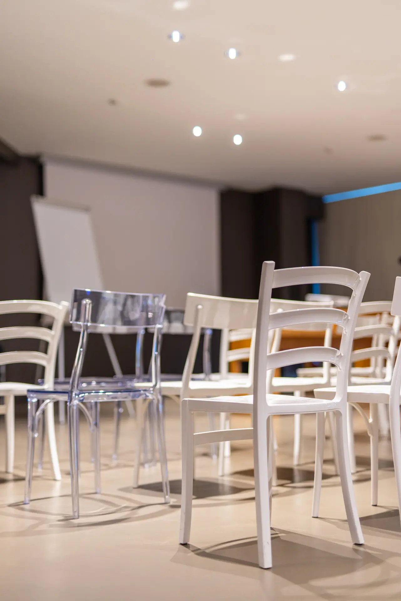 Sedie bianche e trasparenti disposte in una sala conferenze con schermo di proiezione sullo sfondo e luci a soffitto.